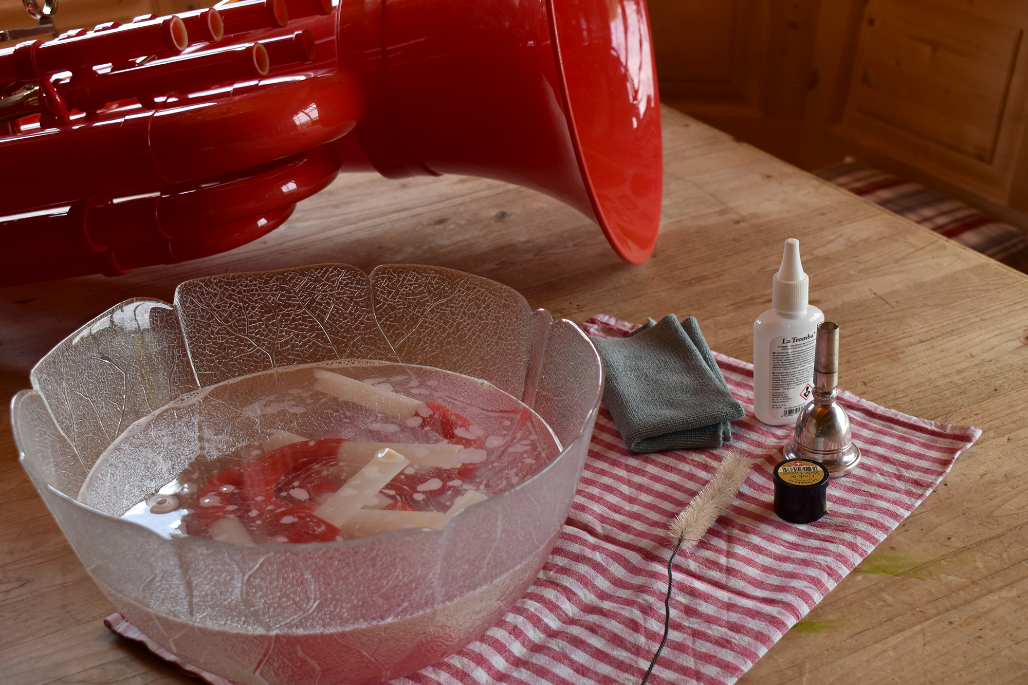 Reinigen der Stimmzüge der Classic Cantabile KT-30RD MardiBrass Kunststoff Bb-Tuba Rot. Die Stimmzüge liegen in einer Glasschüssel in einem Reinigungsbad. Im Hintergrund sieht man die rote Tuba auf einem Holztisch liegen. Neben der Glasschüssel mit dem Reinigungsbad befinden sich mehrere Reinigungs- und Pflegeutensilien.